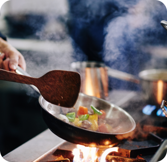 Close-up of vegetables being stir-fried in a wok over an open flame, with steam rising from the cooking surface.