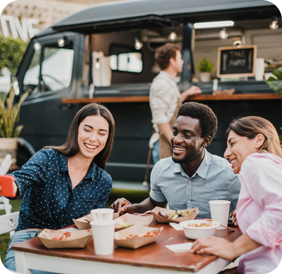 Three customers seated at an outdoor table with meals and drinks, dining beside a food truck with an open service window.