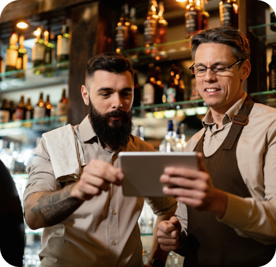 Two apron-wearing staff members viewing a tablet inside a restaurant with illuminated shelves and beverage displays.