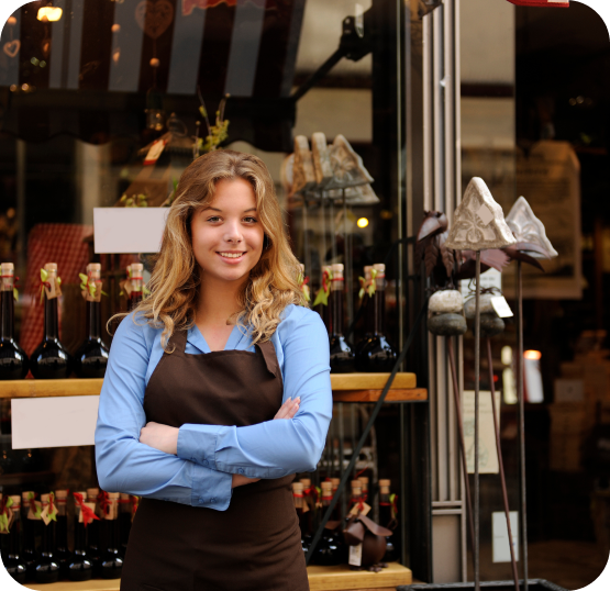 Person wearing a blue shirt and dark apron standing outside a storefront with bottle displays and decorative shelving.