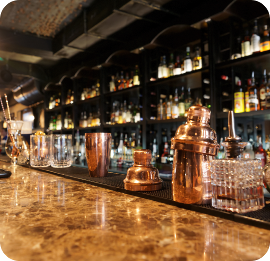 Bar counter displaying cocktail shakers, glassware, beverage tools, and liquor bottles arranged on shelving in the background.