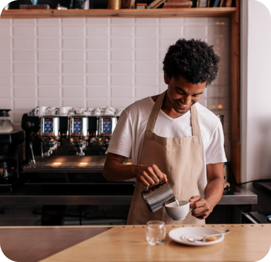 Cafe employee pouring milk into a coffee beverage at a café counter with espresso machines in the background.